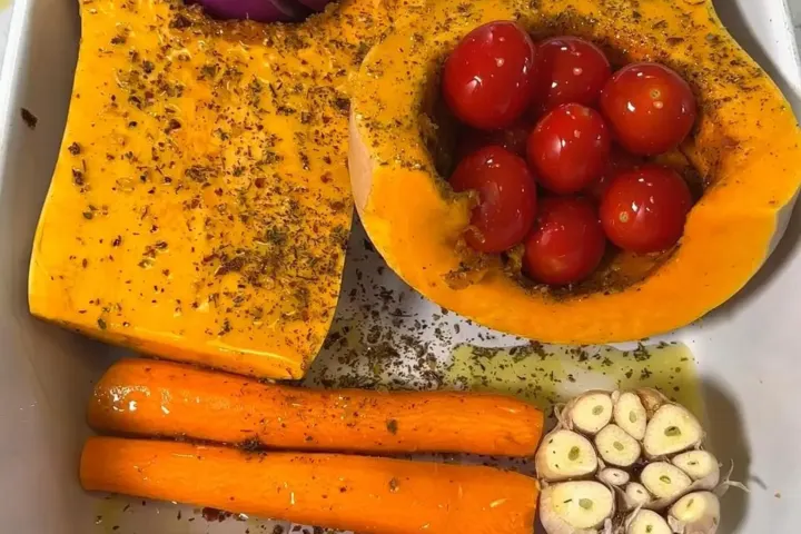Raw butternut squash, carrots, cherry tomatoes, red onion, and garlic seasoned in a baking dish before roasting for soup